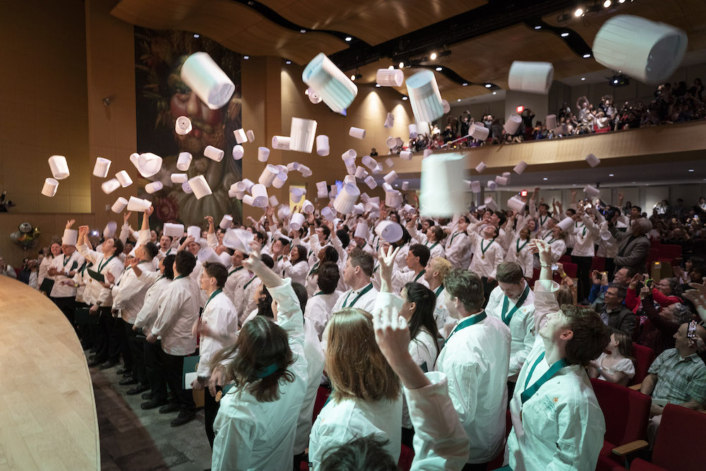 Graduating CIA students celebrate by throwing their chef's hats into the air as their friends and family take pictures.