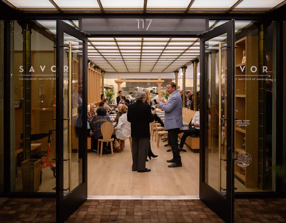 Street view of the CIA's Savor restaurant in San Antonio. The restaurant is full of patrons who are eating, drinking, and talking.