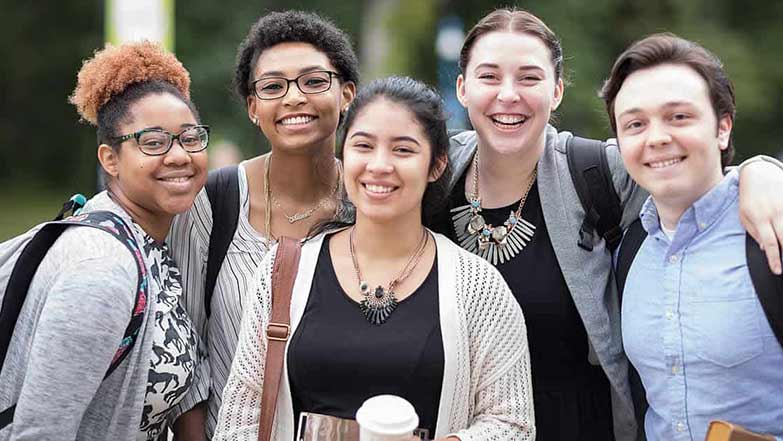 A group of five diverse young adults stands closely together, smiling warmly at the camera. They appear to be students, each carrying backpacks and wearing casual clothing. Trees and greenery are visible in the blurred background, suggesting an outdoor setting.