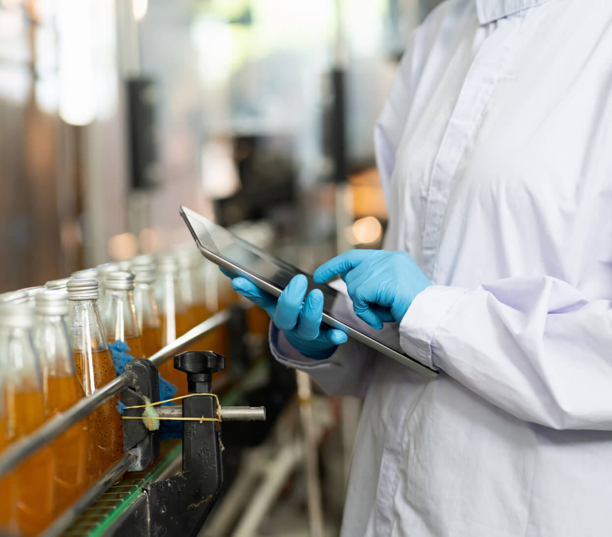 A person wearing a white lab coat and blue gloves is using a tablet while standing next to an assembly line of glass bottles filled with a yellowish liquid. The clean, industrial production environment suggests an emphasis on precision and efficiency, crucial aspects of food service management.