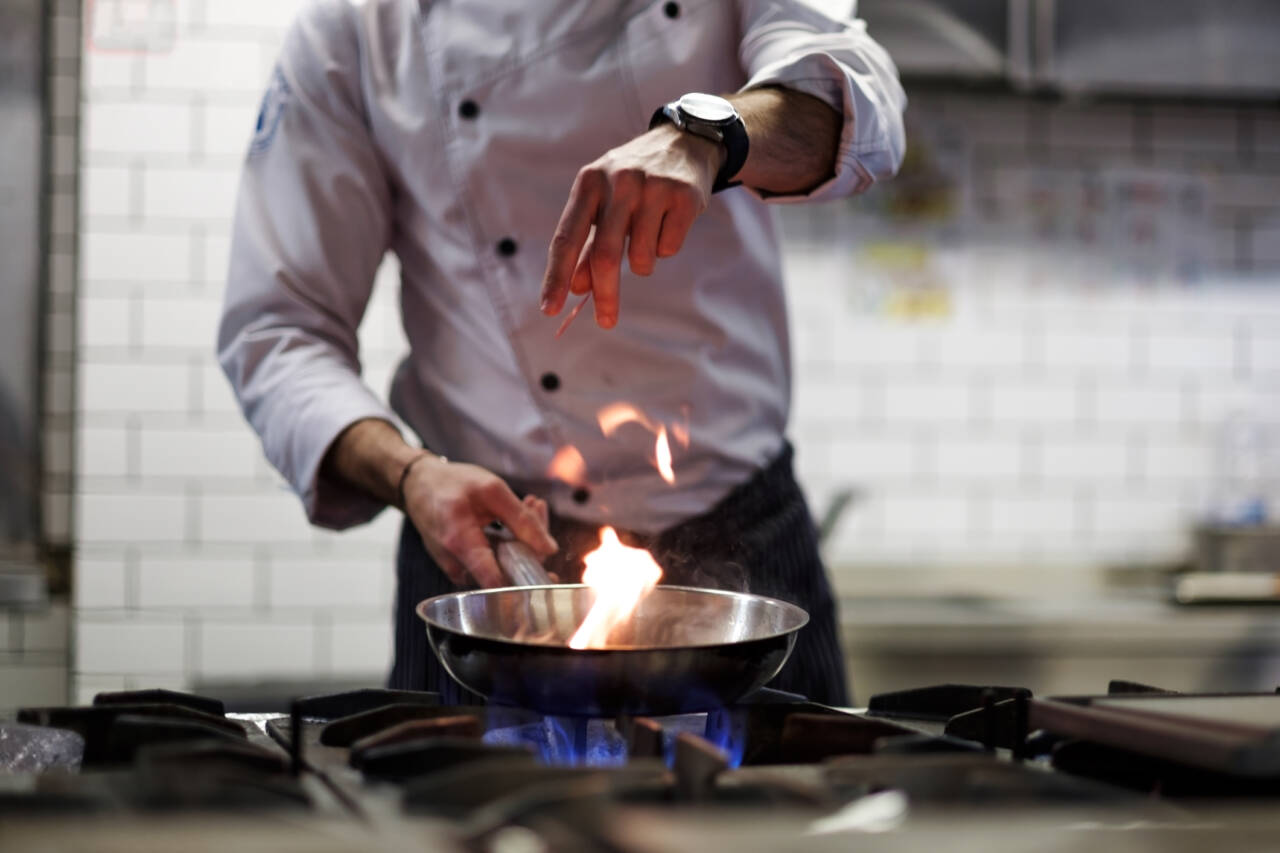 A chef in a white uniform is cooking in a professional kitchen. He is holding a frying pan over a blue gas flame, with a small fire rising from the pan. The background features white tile walls and stainless steel appliances.