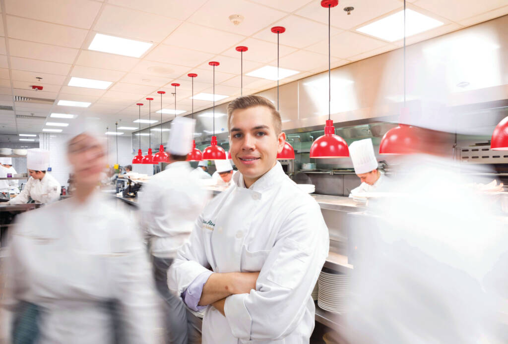 A chef in a white uniform stands confidently with arms crossed in the center of a bustling professional kitchen. Several other chefs, graduates of prestigious culinary arts programs, work diligently in the background, with motion blur highlighting the busy atmosphere. Red heat lamps are above the counters.