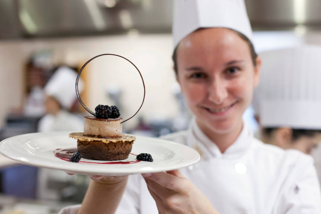 A chef in a white uniform and hat holds up a beautifully plated dessert with a chocolate base, mousse topping, blackberries, and a decorative chocolate ring. The kitchen background is slightly blurred.