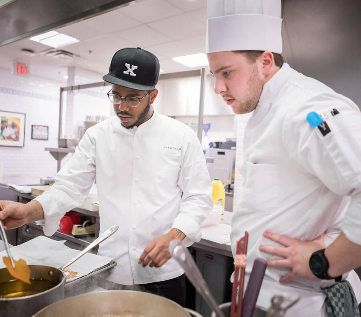 Two chefs working in a kitchen. One chef, wearing glasses, a black cap, and a white jacket, is stirring a pot on the stove. The other chef, wearing a white hat and jacket, is observing him with hands on hips. The kitchen is bright and modern with stainless steel appliances.