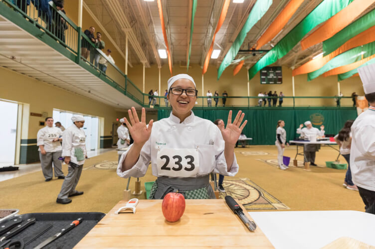 A person wearing a chef's uniform and number 233 bib stands behind a table with a cutting board and an apple, smiling and holding up both hands. The indoor setting at the CIA Hyde Park Campus includes other chefs and green and orange ceiling decorations.