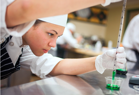 A focused chef wearing a white uniform and hat uses a pipette to carefully measure green liquid into a small plastic container. The chef is in a professional kitchen with other chefs working in the background.