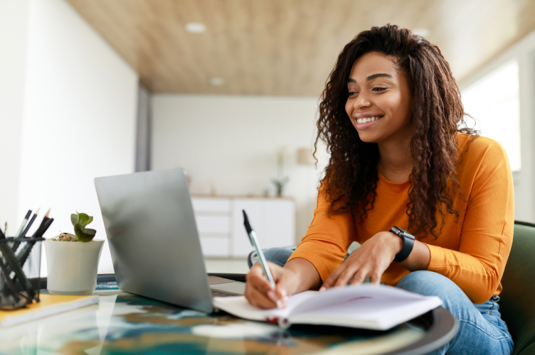A person with long curly hair is sitting at a table, smiling while working on a laptop and writing in a notebook. They are wearing an orange long-sleeve shirt and a smartwatch, and appear to be in a bright, modern room with minimal decor.