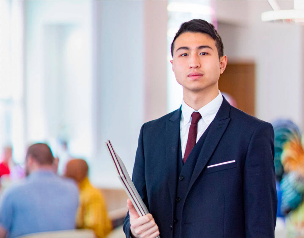 A person dressed in a sharp navy-blue suit with a burgundy tie stands confidently indoors, holding a notebook and a pen. The background is softly blurred, showing other individuals and a vibrant, colorful mural.