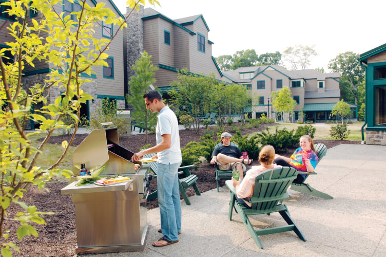 A person is grilling food on a barbecue in an outdoor area near a set of green lawn chairs. Three people are sitting in the chairs, relaxing and chatting. The setting appears to be part of the cia hyde park campus, complete with multi-story buildings and landscaped gardens.