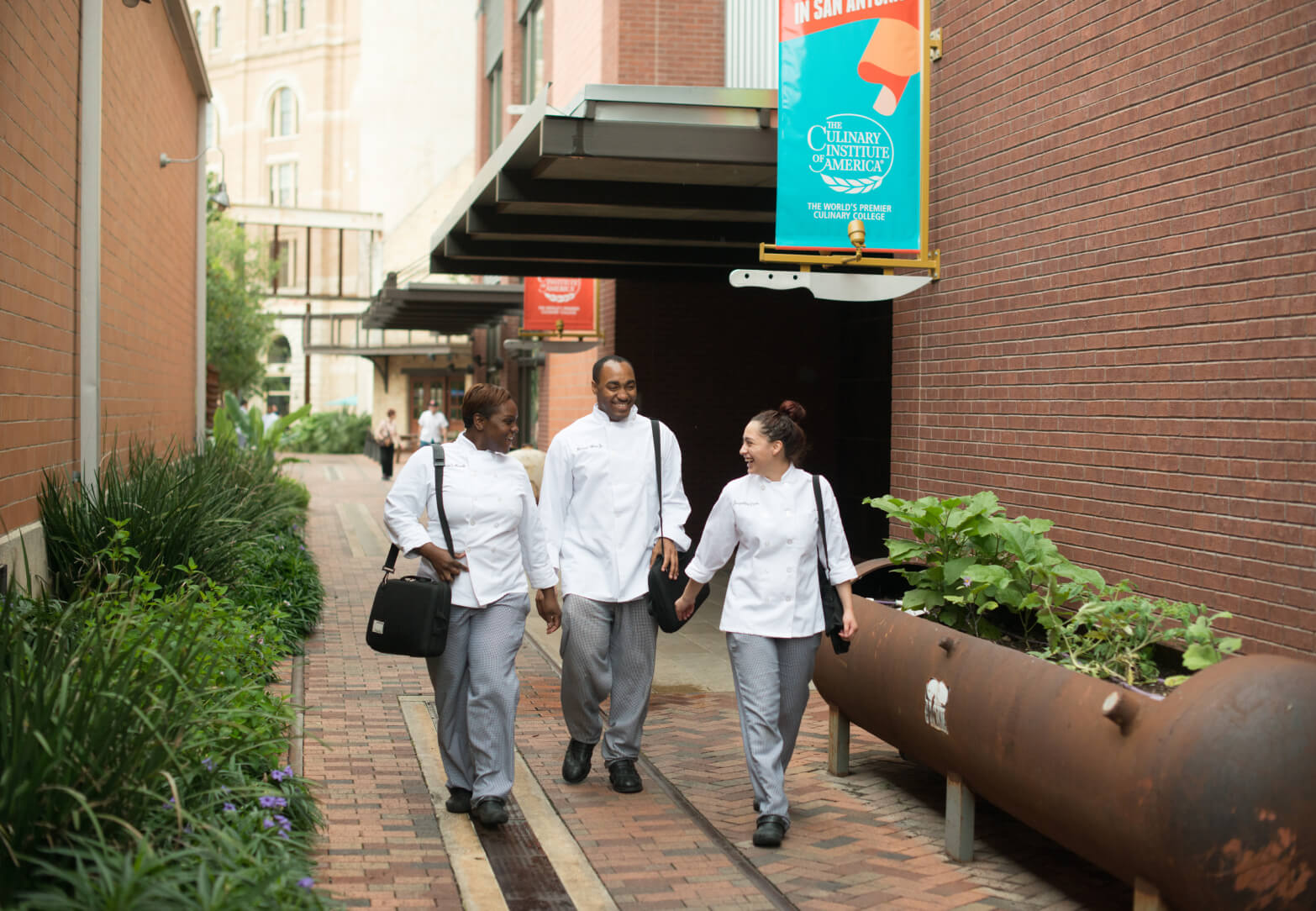 Three people in chef uniforms walk together down a brick pathway between buildings. They are talking and smiling. One person carries a black bag. There is a sign overhead that reads 