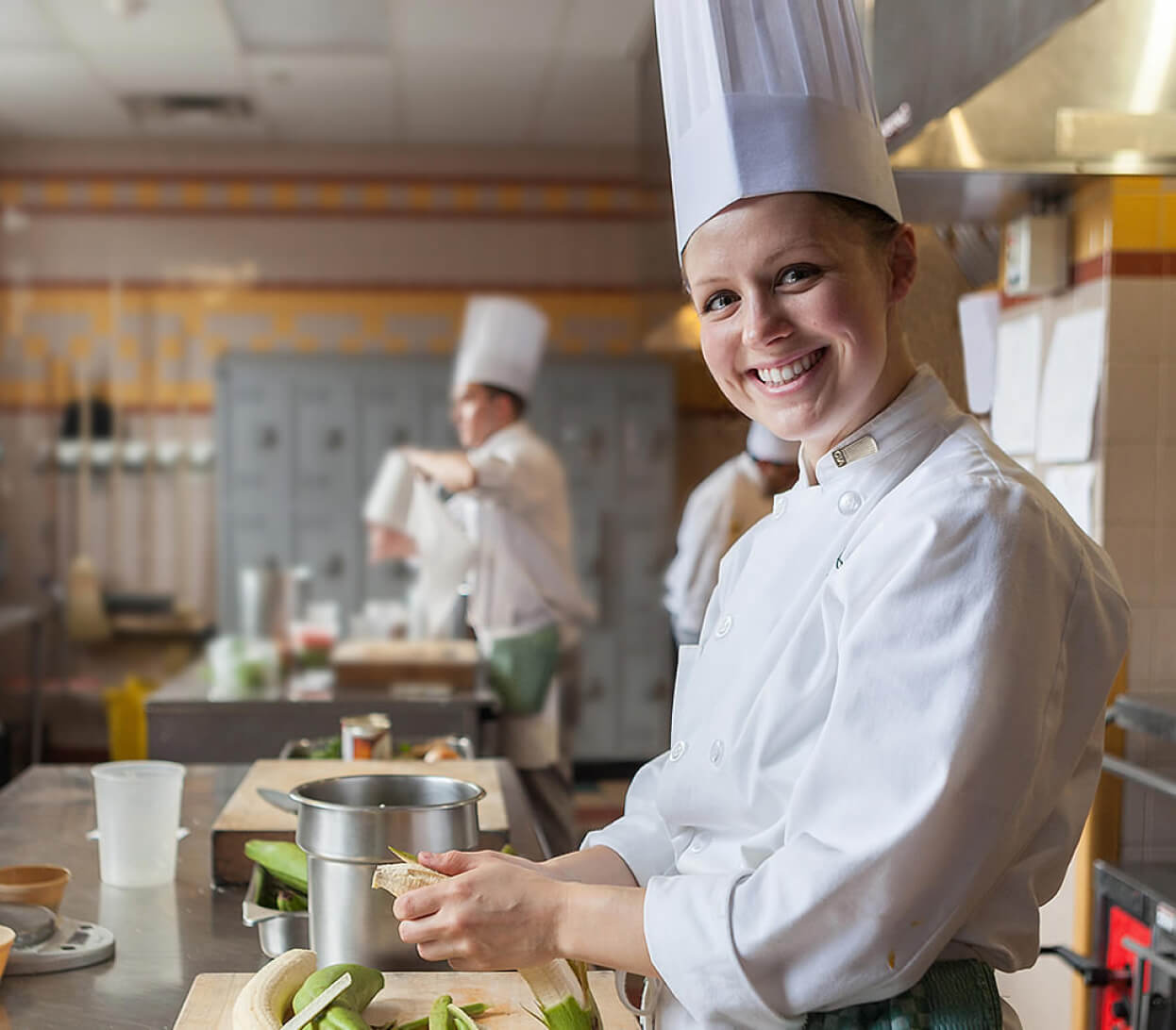 A smiling chef wearing a tall white hat and white uniform is standing at a kitchen counter, peeling vegetables. In the background, other chefs in similar attire are busy cooking. The kitchen has a clean, organized appearance with stainless steel surfaces.