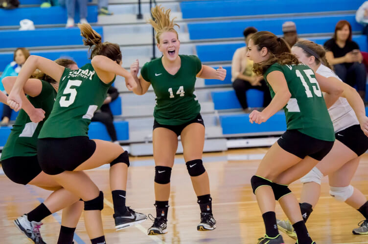 A group of female volleyball players wearing green jerseys and black shorts celebrate on the court at the CIA Hyde Park campus. The player with jersey number 14 appears to be shouting with joy. They are in an indoor gymnasium with blue bleachers and spectators in the background.