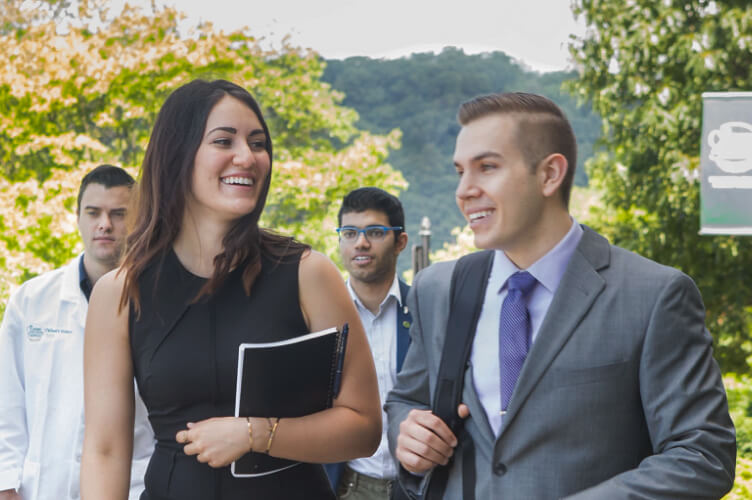 A group of people walking outdoors on a sunny day at the Hyde Park campus. A woman in a black dress and a man in a gray suit, possibly CIA agents, are smiling and talking to each other while others follow behind them, including a person in a white lab coat and another in casual attire. Trees and hills are in the background.
