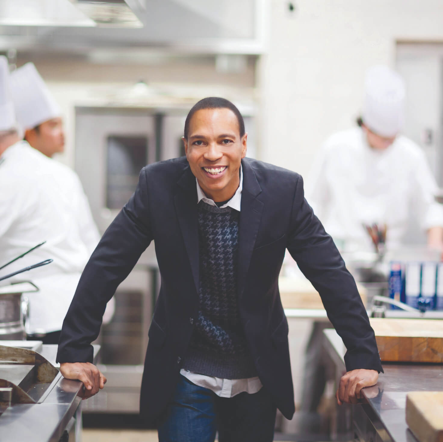 A man in a professional kitchen smiles at the camera. He is wearing a dark blazer over a sweater and collared shirt. <a href=