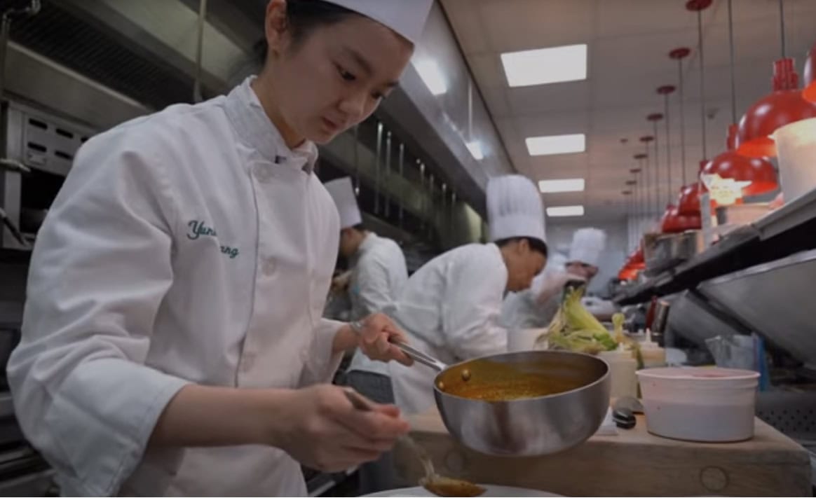 A chef in a white uniform and hat is carefully plating a dish with sauce from a pot in a busy professional kitchen. Other chefs in white uniforms and hats are actively working in the background, under red heat lamps.