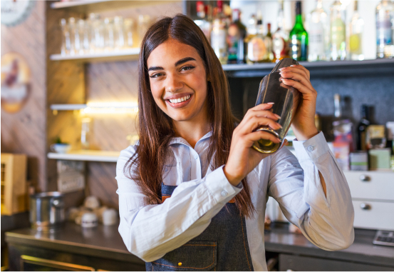 A smiling woman with long brown hair, wearing a light blue shirt and a dark apron, is shaking a cocktail shaker behind a bar counter. The background features various bottles and bar supplies on shelves.