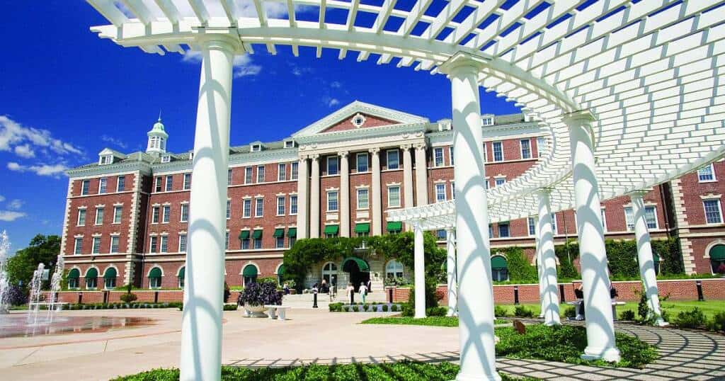 A grand, multi-story brick building with white columns and green window awnings stands under a clear blue sky. In the foreground, a white pergola with curved beams and supporting columns creates a geometric pattern. Fountains and greenery adorn the plaza in front.