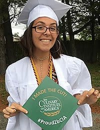 A graduate, wearing a white gown and cap, is smiling and holding her decorated graduation cap. The cap reads "I MADE THE CUT!" along with "The Culinary Institute of America" and the hashtag "#Proud2bCIA".