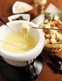 A fondue pot filled with melted cheese is surrounded by skewers holding bread cubes. In the background, there are bowls of cubed bread, pickles, and a pepper grinder. The scene is set on a wooden table.