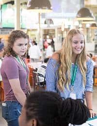 Two young girls are standing indoors, engaging in a lively environment filled with people. The girl on the right has long blonde hair and is wearing a striped off-shoulder top, while the girl on the left has curly hair and is wearing a purple shirt. Both have lanyards around their necks.