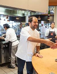 A chef wearing a white coat and shaking hands with someone off-camera in a busy kitchen. Other kitchen staff members in the background are preparing food and working at various stations. The kitchen has stainless steel appliances and cabinets with a warm atmosphere.