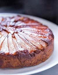A close-up of a round caramelized pear upside-down cake on a white plate. The pears are thinly sliced and arranged in a circular pattern on top of the golden-brown, slightly crispy cake. The cake has a glossy finish, indicating it is very moist.