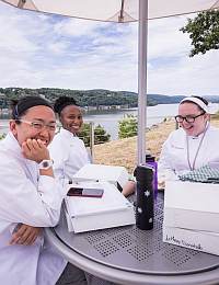 Three people in lab coats sit at an outdoor table near a body of water, smiling and having a discussion. The background features trees and hills under a partly cloudy sky. Several items, including a phone, water bottle, and boxed meals, are on the table.