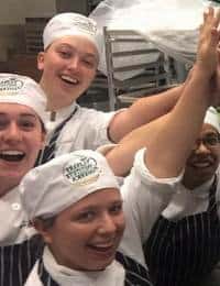 Four individuals wearing chef uniforms and hats are happily raising their arms in celebration in a kitchen. They appear to be in good spirits and standing in front of a shelf with metal trays.