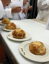 Several rounds of bread or pastry on plates labeled "Brioche Fine" are displayed on a white countertop. People in white chef coats are seen in the background examining and discussing the baked goods.