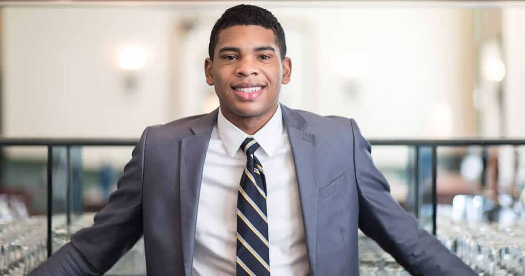 A young man in a gray suit and striped tie smiles confidently while standing with his arms resting on a glass counter. The background features a blurred interior with warm lighting.
