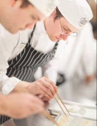 Two chefs in white uniforms and hats focus intently on their task in a bright kitchen. One chef in the foreground uses tongs to position food, while the other chef in the background is absorbed in a similar culinary activity.