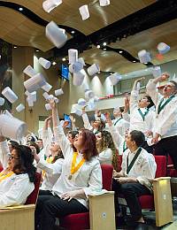 A group of people dressed in white coats and wearing honor cords are celebrating by tossing rolled-up papers into the air. They are seated in an auditorium with red chairs and a wooden ceiling, creating a festive and jubilant atmosphere.