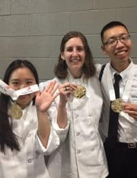 Three people wearing white chef coats are standing together, each displaying a gold medal around their necks. The person on the left is holding their medal with their teeth and waving, the person in the middle is smiling, and the person on the right is also smiling widely.