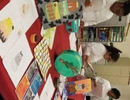 A display table with various art pieces, including paintings, drawings, and a decorated cake. Three people in white uniforms are standing behind the table examining some of the artworks. Shelves with books and supplies are visible in the background.