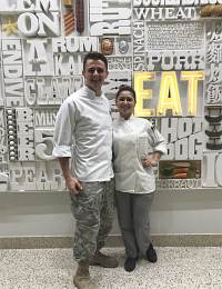 Two chefs stand side by side in front of a wall decorated with various food-related words and an illuminated "EAT" sign. The man on the left wears a white chef's coat and camouflage pants, while the woman on the right wears a white chef's coat and gray pants. They are both smiling.