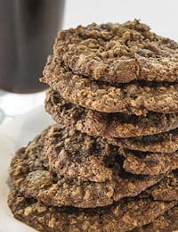 A close-up image of a stack of oatmeal cookies on a plate. The cookies are textured with visible oats and appear chewy. In the background, there's a blurry glass of milk.