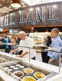 People selecting food at a self-service buffet counter with a prominent sign reading "THE LINE" in the background. The counter features various dishes in metal trays, and the area is bustling with activity.