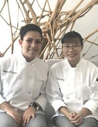Two chefs wearing white uniforms sit side by side, smiling at the camera. They are seated in front of a background with intertwined wooden branches, providing a natural and rustic decor element.