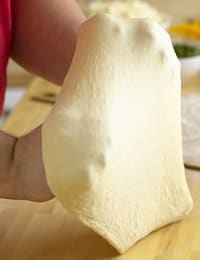 A person stretches a piece of pizza dough with both hands over a wooden surface. The dough appears smooth and elastic, showing some translucency. In the background, blurry ingredients and kitchen items can be seen.