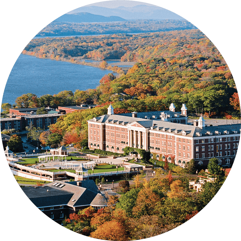 Aerial view of a large, picturesque campus featuring a prominent, stately brick building with columns at the entrance. The campus, reminiscent of Culinary Institute of America locations, is surrounded by lush autumn trees and is adjacent to a large river, with a backdrop of distant hills.