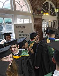 A group of students dressed in black graduation caps and gowns gather in a hallway with a brick wall and large windows. Some have colorful tassels and hoods. A sign in the background reads, "Ask a Bachelor's Graduate." The atmosphere is celebratory.