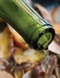 A close-up of a green glass bottle pouring liquid, probably wine, over a dish of clams. The image focuses on the bottle's mouth and the stream of liquid emerging from it, with a blurred background of clamshells.