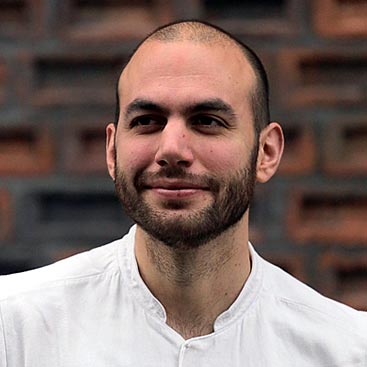 A man with a shaved head and trimmed beard smiles while looking slightly to the left. He is dressed in a white shirt and stands in front of a dark, textured brick wall background.