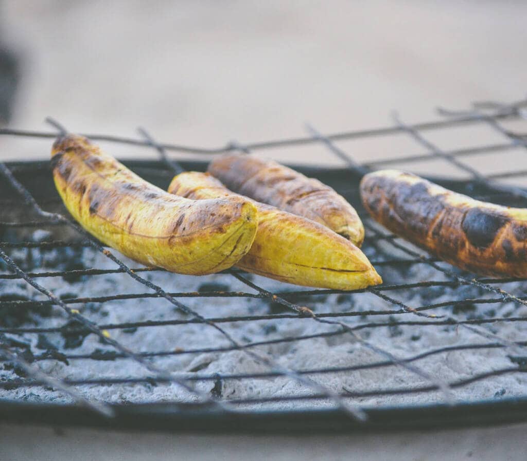 Four ripe plantains grilling on an outdoor barbecue, with char marks visible on the skins. The grill grate is slightly worn, and the background is blurred.