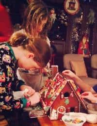 Two children are decorating a gingerbread house at a table. The child with blonde hair is standing in the back, while the child with reddish-brown hair and wearing an owl-patterned shirt is in the foreground. The room is festively decorated for the holidays.