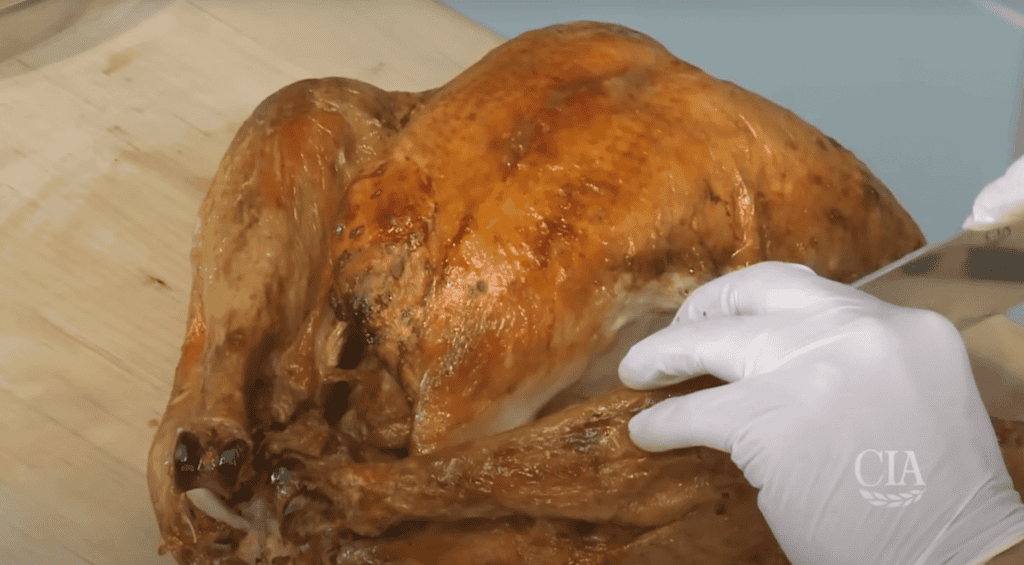 A person wearing white gloves is carving a roasted turkey with a knife on a wooden cutting board. The golden-brown roasted turkey is being sliced, revealing the white meat inside. There is a partial view of a glass bowl in the background.