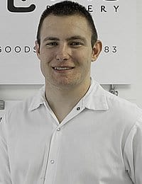 A man with short brown hair and a slight smile is pictured wearing a white collared shirt. He stands in front of a white wall with partially visible text.