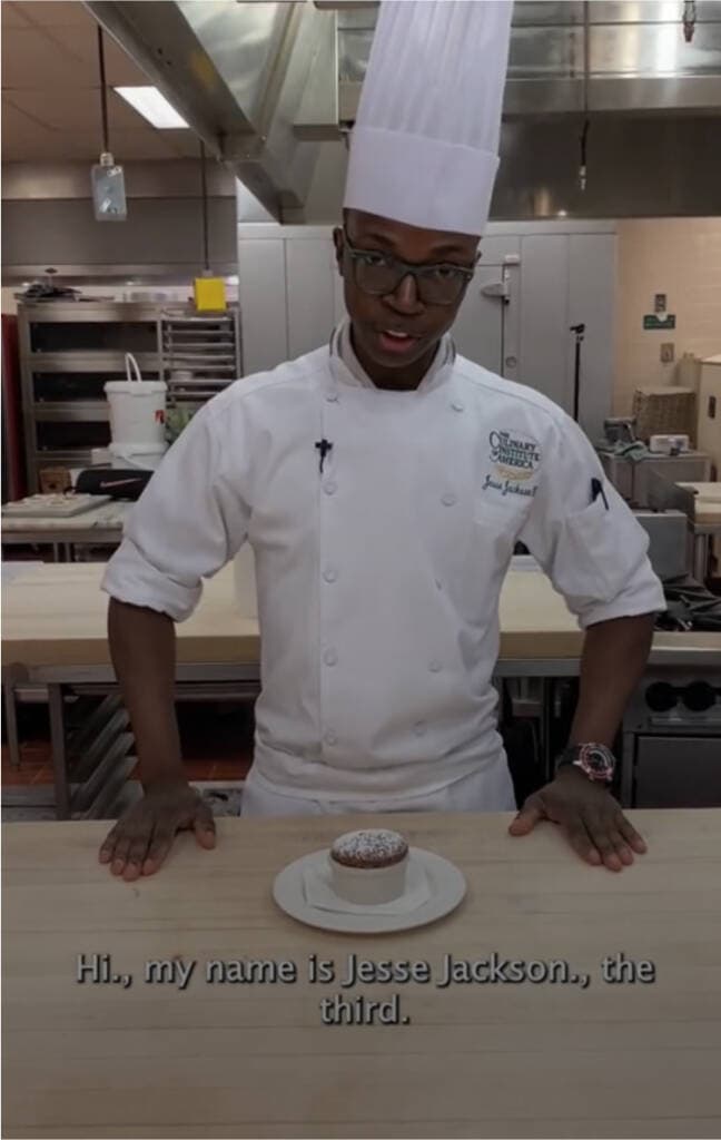 A chef in a white coat and tall hat stands in a professional kitchen, smiling at the camera. In front of him on the counter is a plate with a dessert. Subtitles read, "Hi, my name is Jesse Jackson, the third.