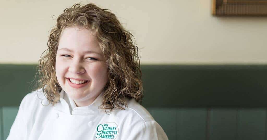 A smiling individual with curly hair is wearing a white chef's coat with "The Culinary Institute of America" logo. They are seated indoors against a green cushioned bench, with light-colored walls in the background.