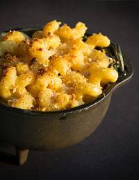 A small cast iron pot filled with baked macaroni and cheese, topped with a golden-brown layer of breadcrumbs. The mac and cheese appears creamy and rich, with a slightly crispy and browned crust.